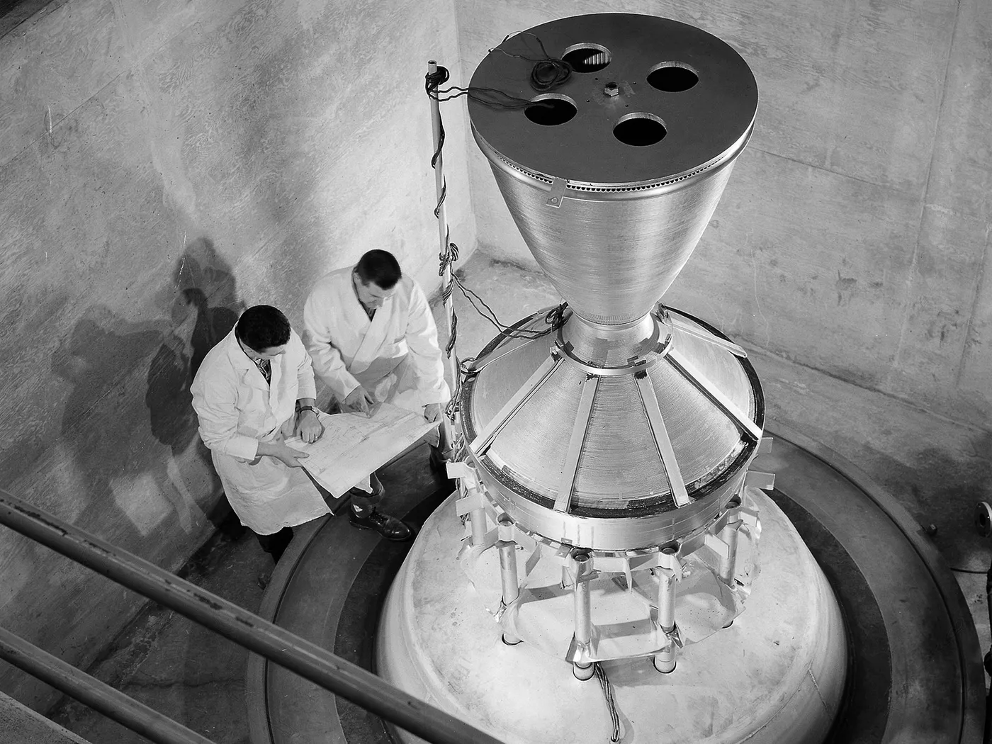 Two technicians in white coats study drawings beside a large conical rocket nozzle assembly in a round concrete pit